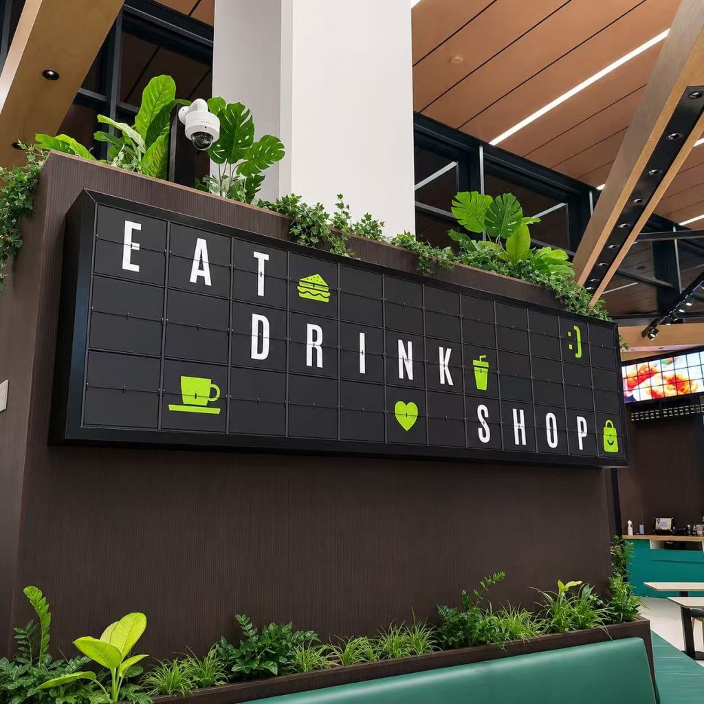 Custom split flap display wall for a restaurant interior showing EAT, DRINK, and SHOP with green icon graphics.