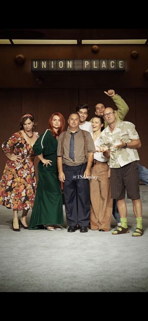 Theatre cast in front of a split flap display showing Union Place