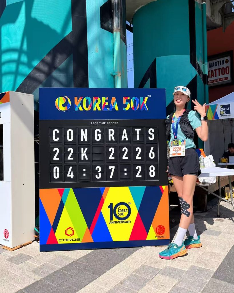 Female race finisher standing beside the colorful KOREA 50K split flap race time display.