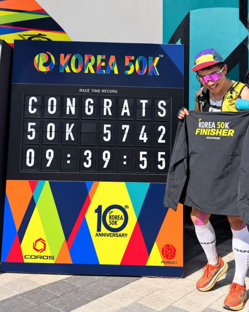 KOREA 50K finisher holding a finisher shirt beside a TS Display split flap race time board.