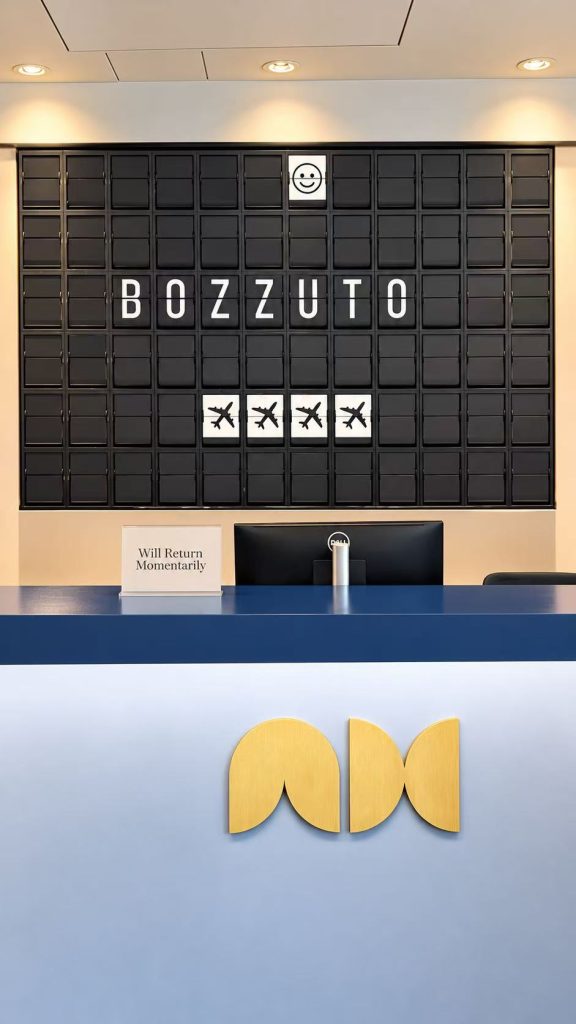 Black split flap display showing BOZZUTO, a smiley face and four airplane symbols behind the blue concierge desk at Aviator House.