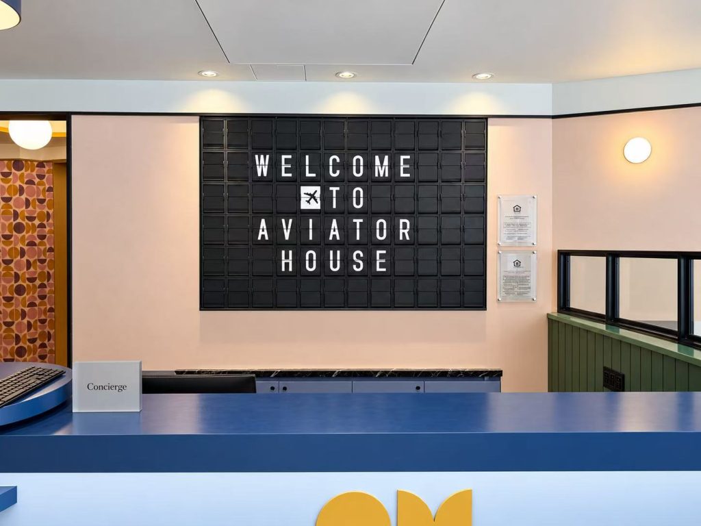 Black split flap display behind a reception desk showing “Welcome to Aviator House” with an airplane icon.