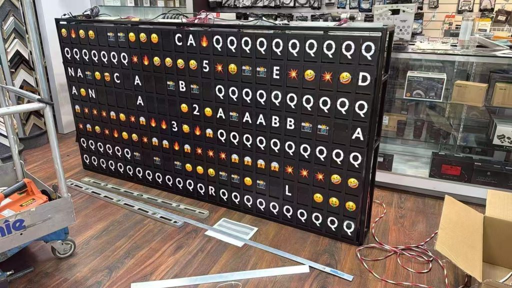 Large black split flap display showing letters, numbers, and emojis on the showroom floor at Unique Photo in Fairfield, New Jersey.
