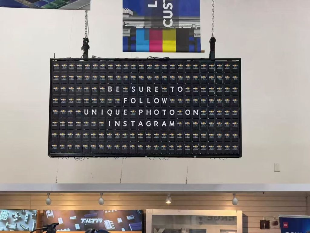 Black split flap display suspended by chains at the Unique Photo showroom in the USA, showing an Instagram follow message and camera icons.
